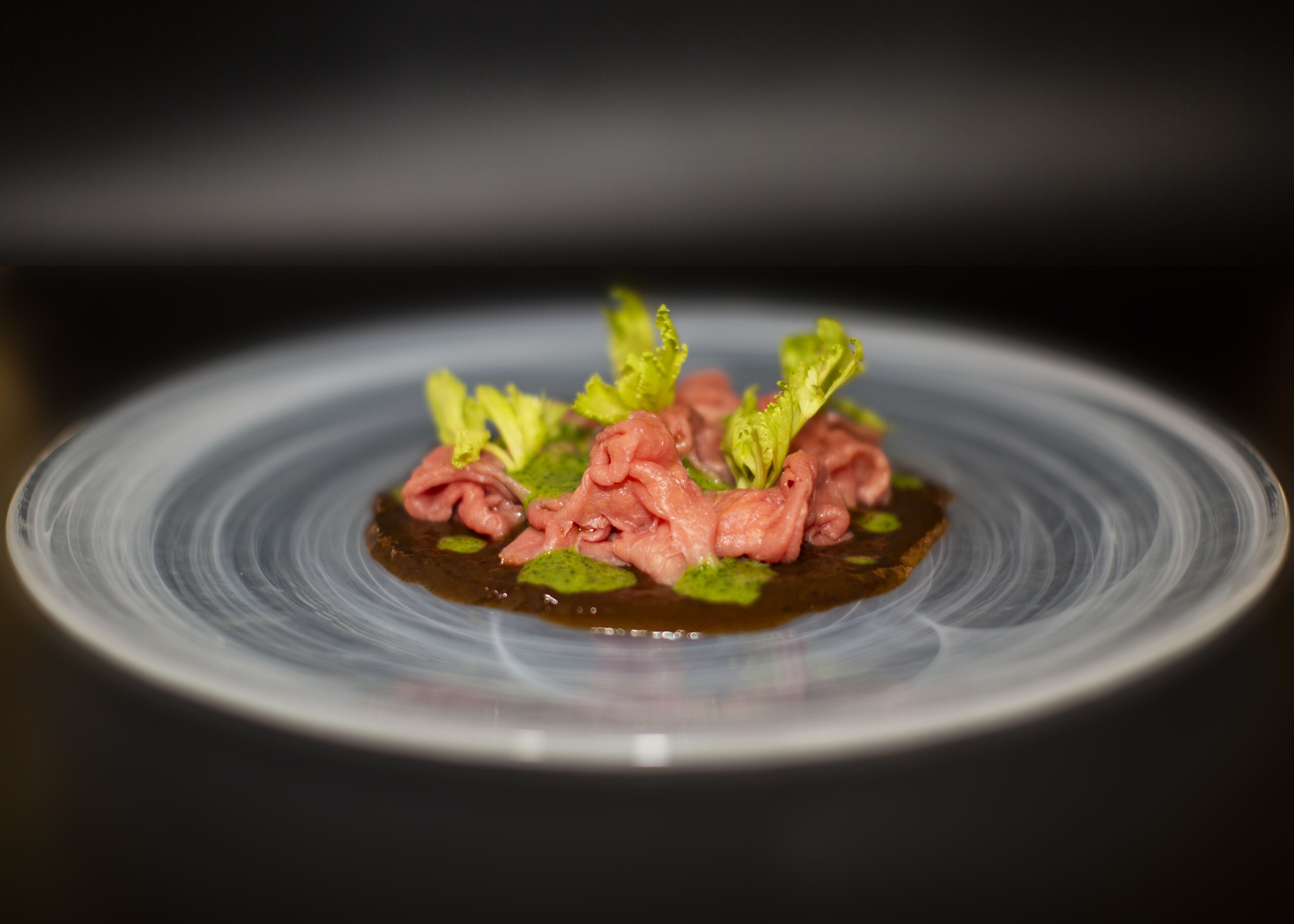 Een bord met donkere saus, roze vlees en groene bladeren.