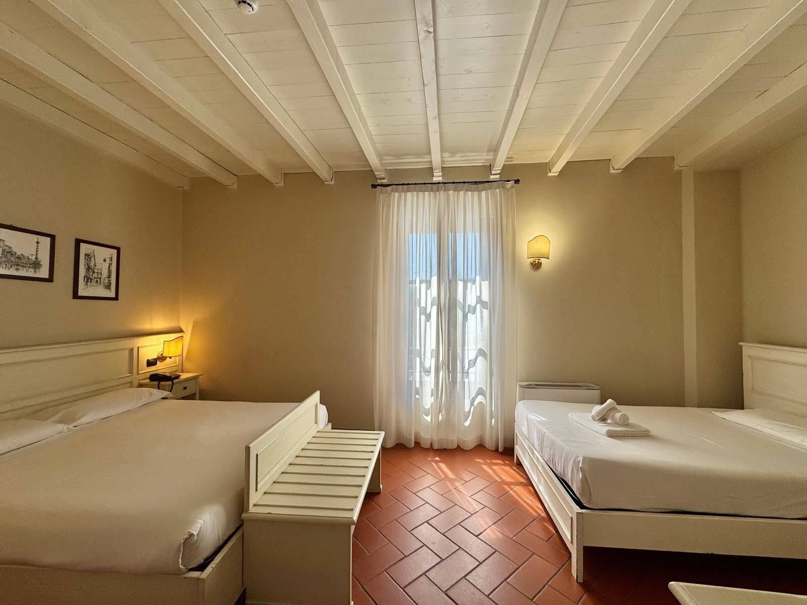 Habitación de hotel con dos camas, una mesita de noche con lámpara, luz empotrada en la pared y ventana con cortinas traslúcidas.