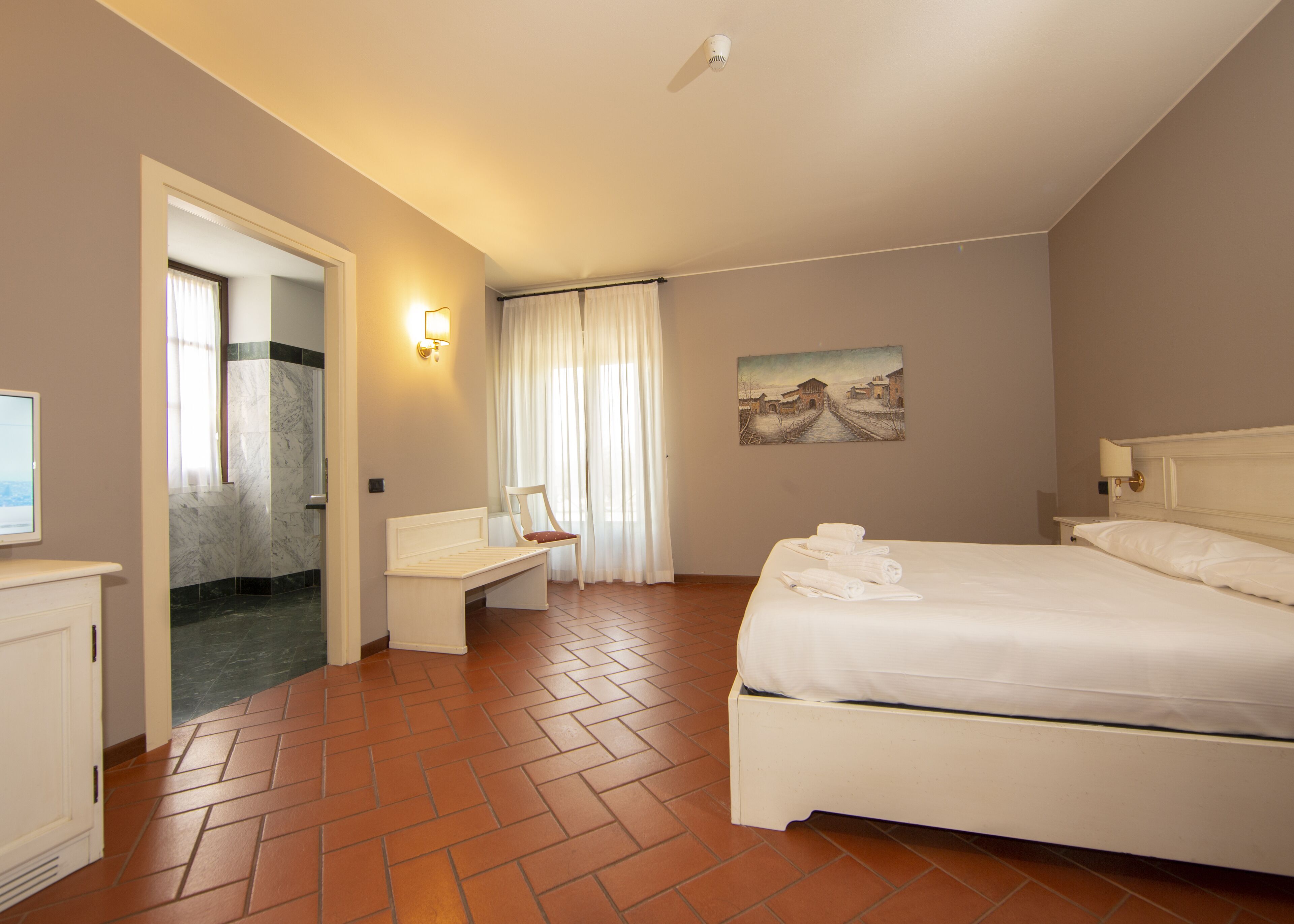 Een hotelkamer met een bed, een bankje, een schilderij en een badkamer met douche en toilet.