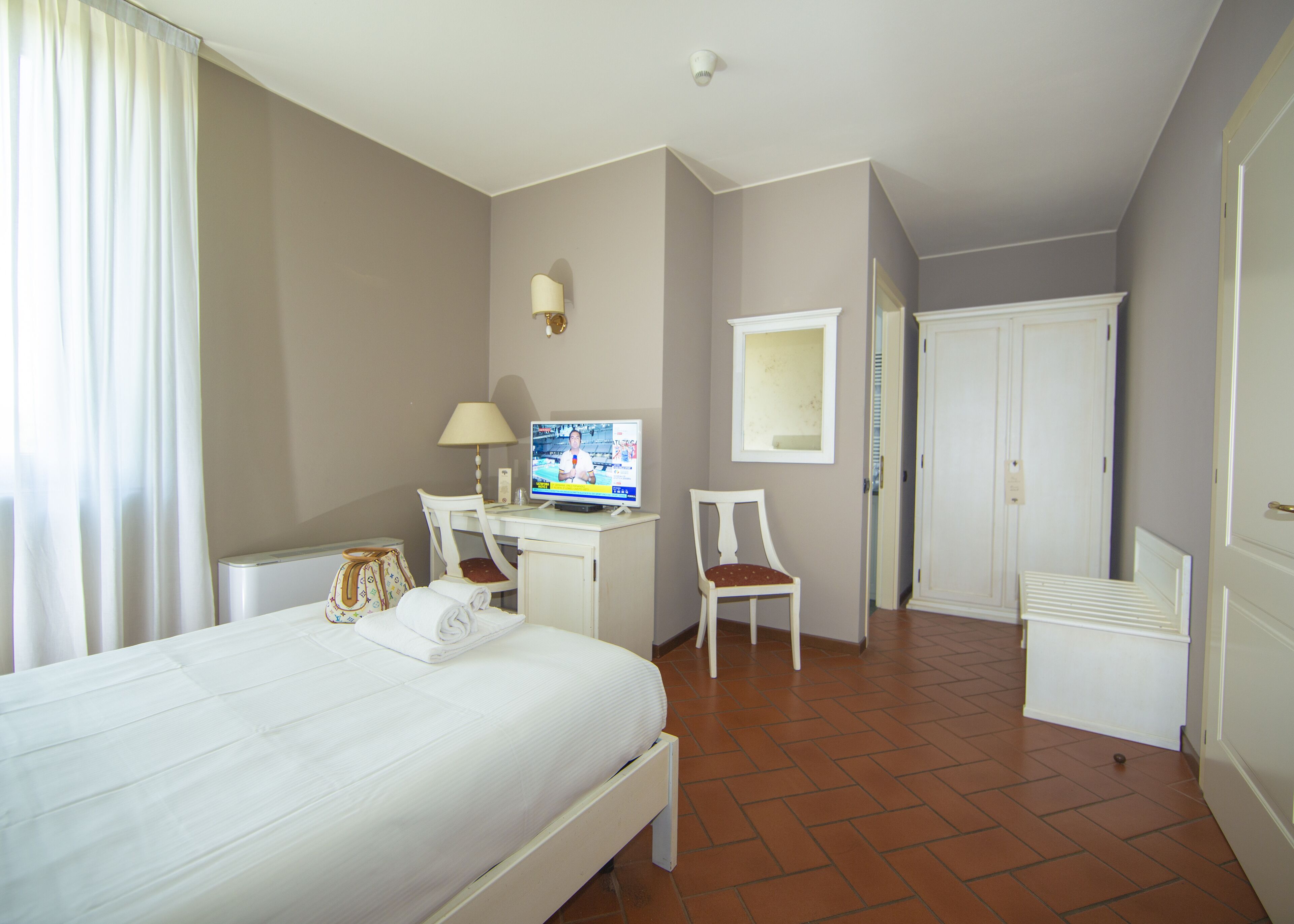 A hotel room with a bed, a desk with a computer, a chair, and a small table with a lamp.