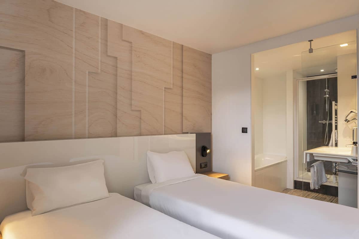 Habitación de hotel con dos camas, cabecero de madera, mesita de noche, baño con ducha y un espejo.