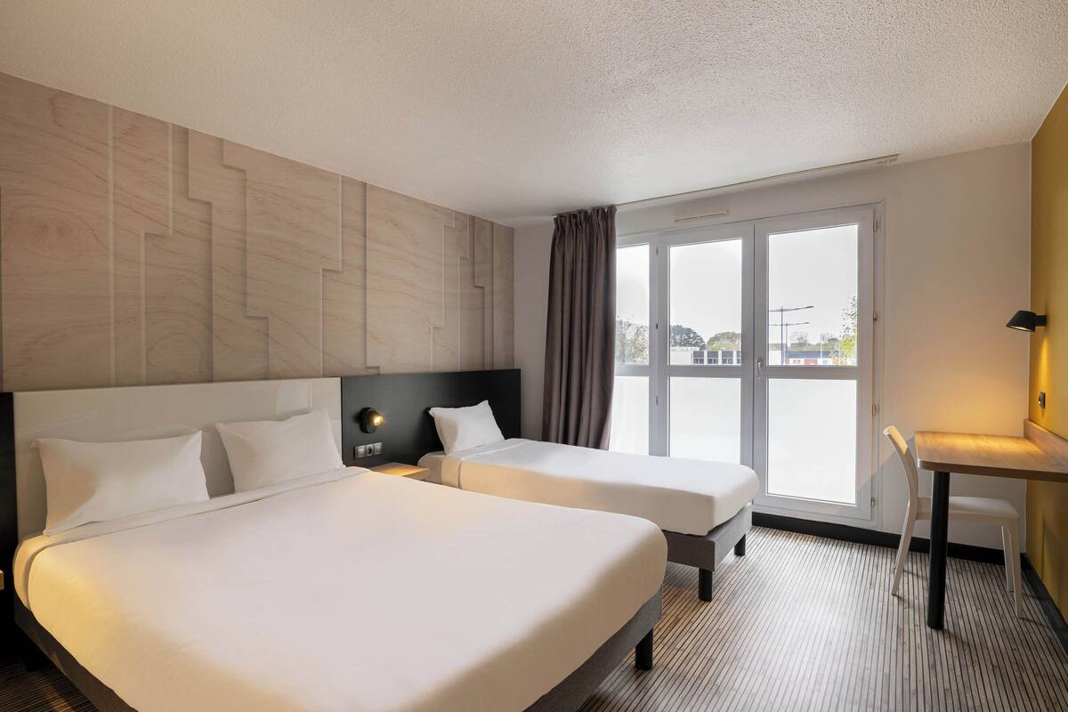 Habitación de hotel con dos camas, un escritorio, una silla y un ventanal con vistas.