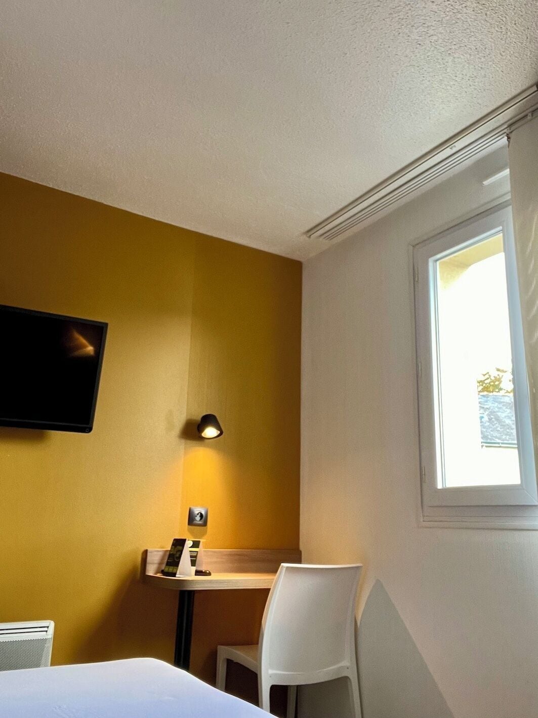 A hotel room with a bed, a desk, a chair, a TV mounted on the wall, and a window.