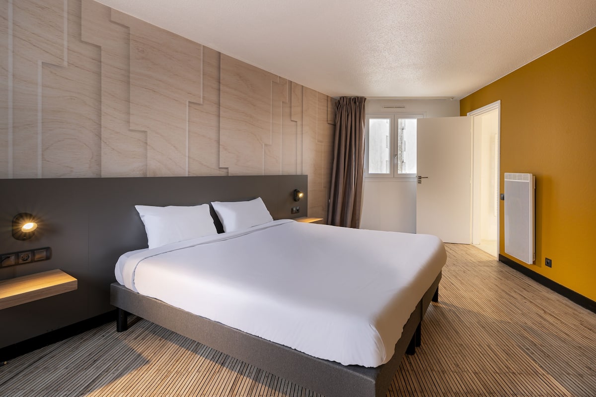 Una habitación de hotel con una cama grande, una mesita de noche con lámpara, cabecera de madera, una ventana con cortinas y un radiador.