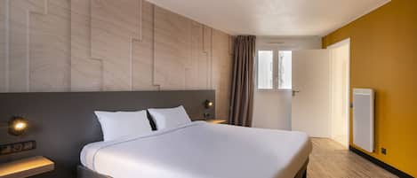 Una habitación de hotel con una cama grande, una mesita de noche con lámpara, cabecera de madera, una ventana con cortinas y un radiador.