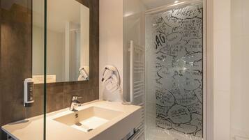 Un baño moderno con un espejo grande, un lavamanos blanco y una cabina de ducha de vidrio con grafiti.