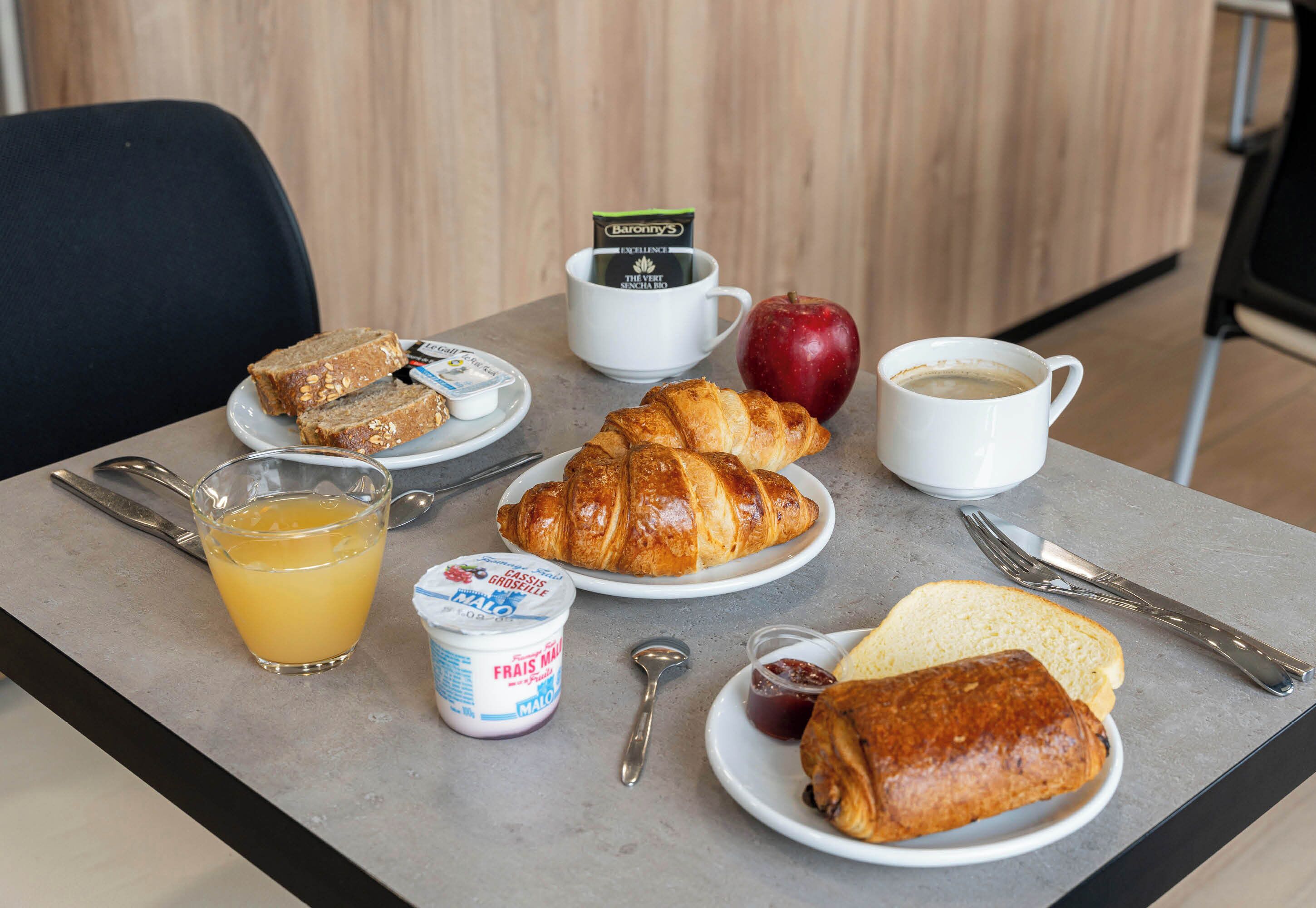 A breakfast table with croissants, bread, an apple, a cup of coffee, a glass of orange juice, and a container of yogurt.