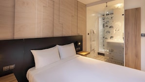 Una habitación de hotel moderna con una cama grande, un cabecero de madera, una mesita de noche y un baño con cabina de ducha de vidrio.
