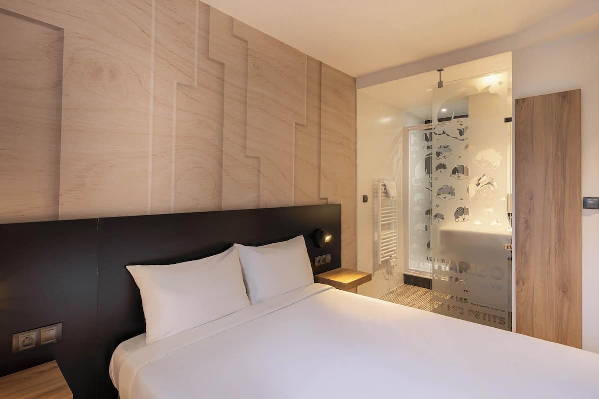 Una habitación de hotel moderna con una cama grande, un cabecero de madera, una mesita de noche y un baño con cabina de ducha de vidrio.