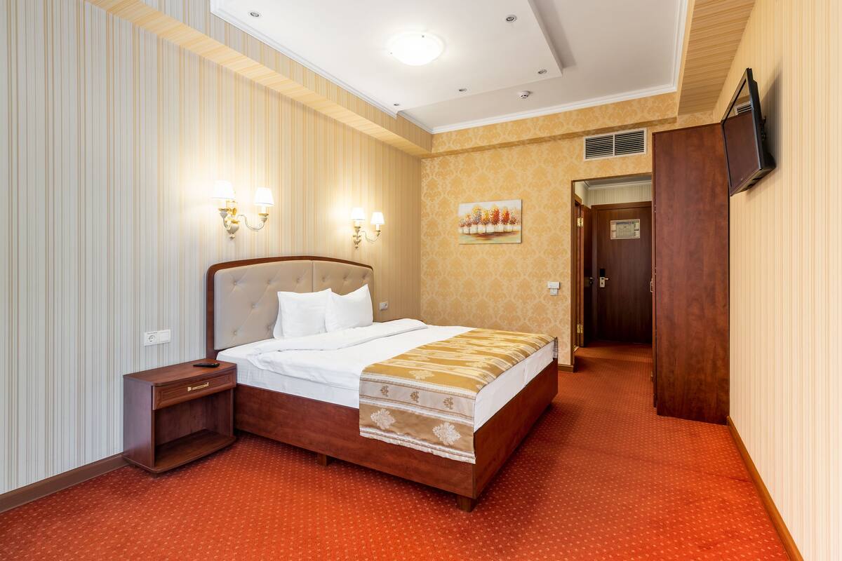 A hotel room with a bed, bedside table, wall-mounted lights, a painting, and a television on the wall.