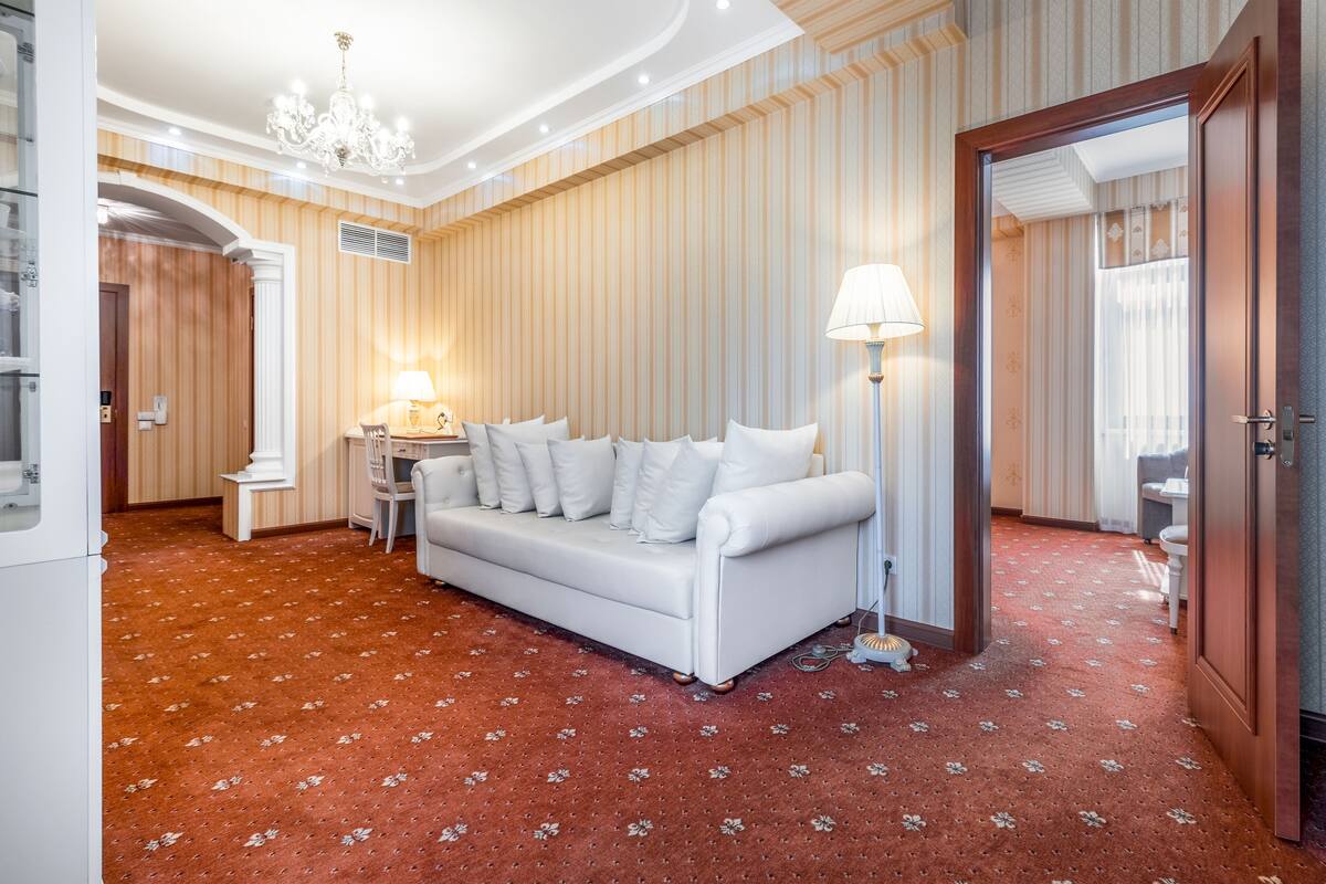 A hotel room with a white sofa, a lamp, a small table, and a red patterned carpet.