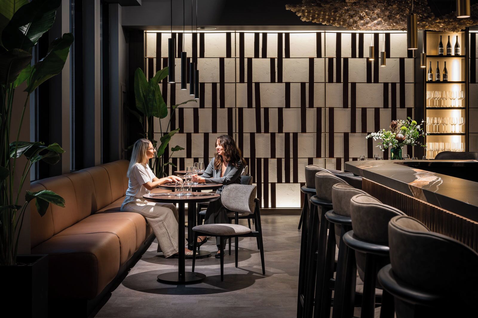 Un área de bar moderna con disposición de asientos, una pared decorativa con paneles verticales y una barra con vasos y botellas.