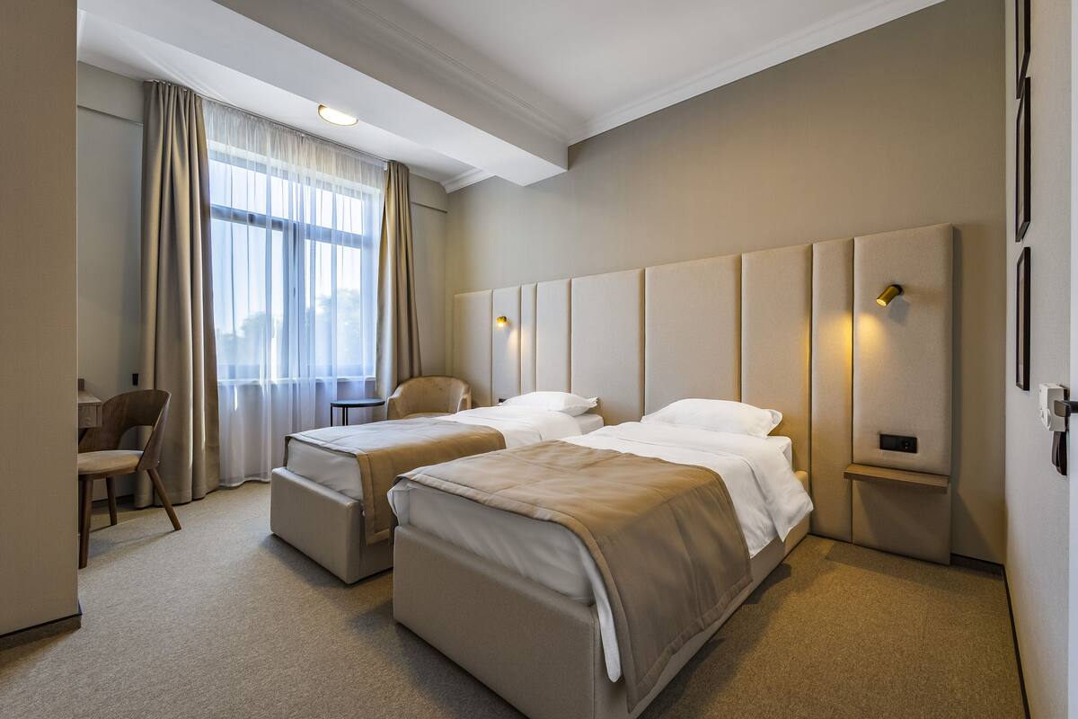 A hotel room with two beds, a desk, a chair, and a large window with curtains.