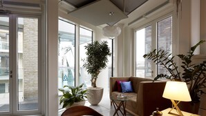 A modern living room with a sofa, a coffee table, and a lamp. There are potted plants and a view of the outside through large windows. - Amadi Park Hotel (Amsterdam)