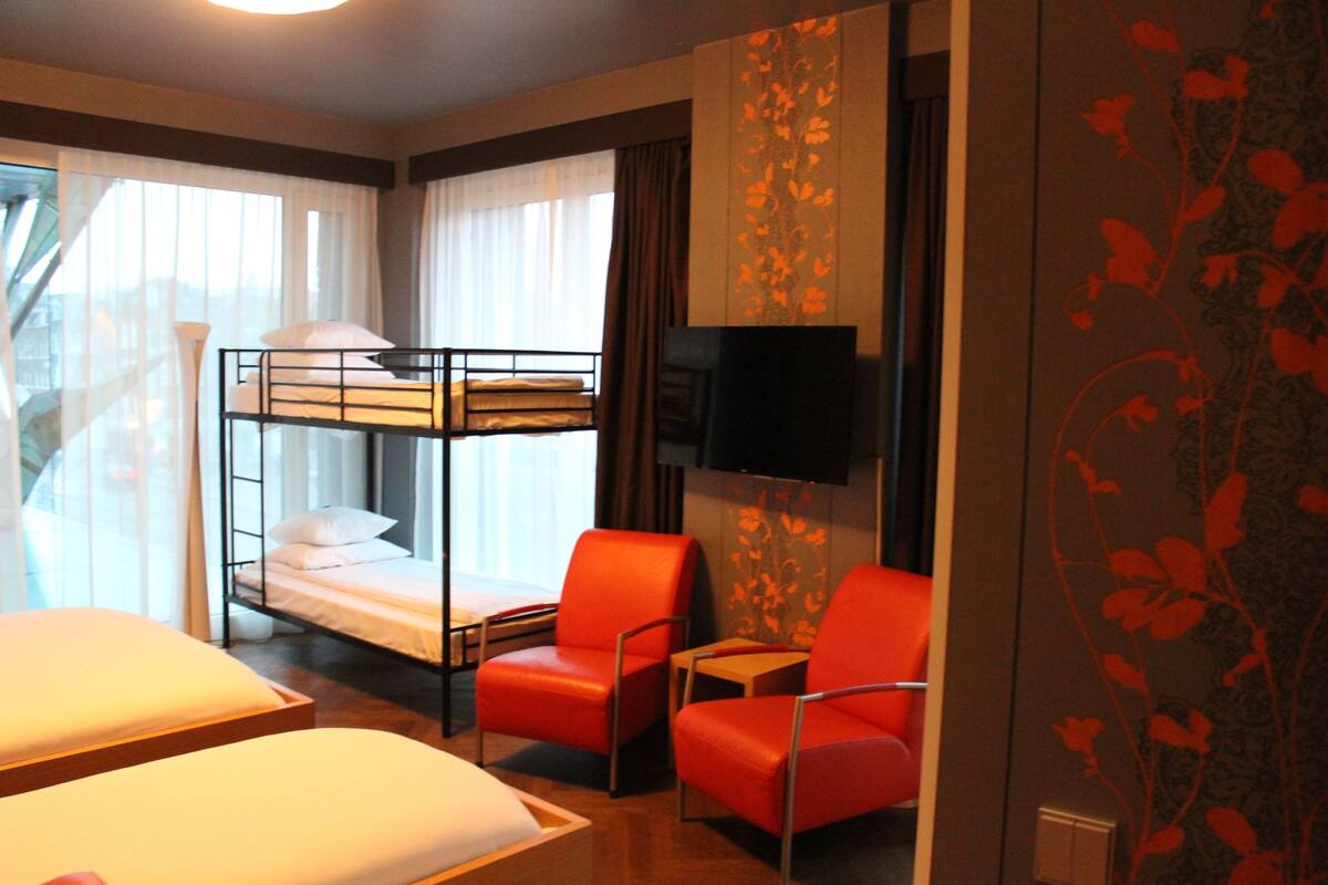 A hotel room with a bunk bed, two red chairs, a flat-screen TV, and large windows with sheer curtains.