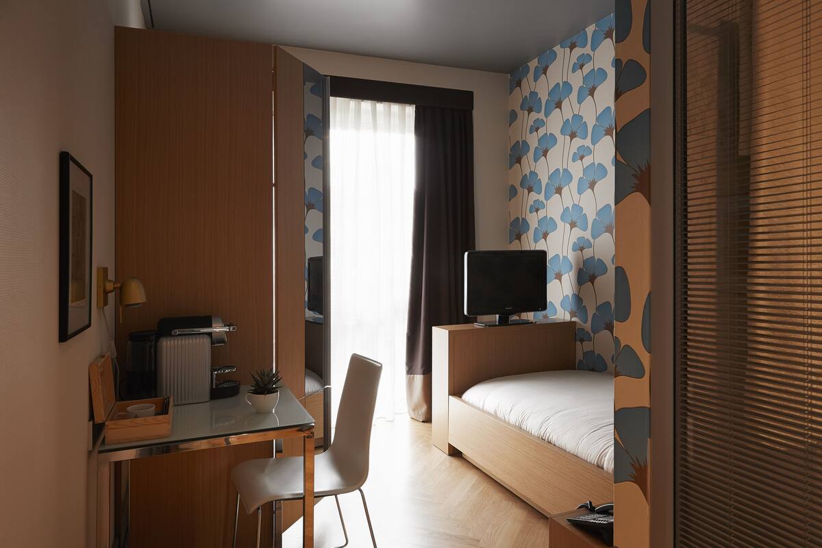 A hotel room with a bed, a desk with a chair, a TV, and a window with curtains.