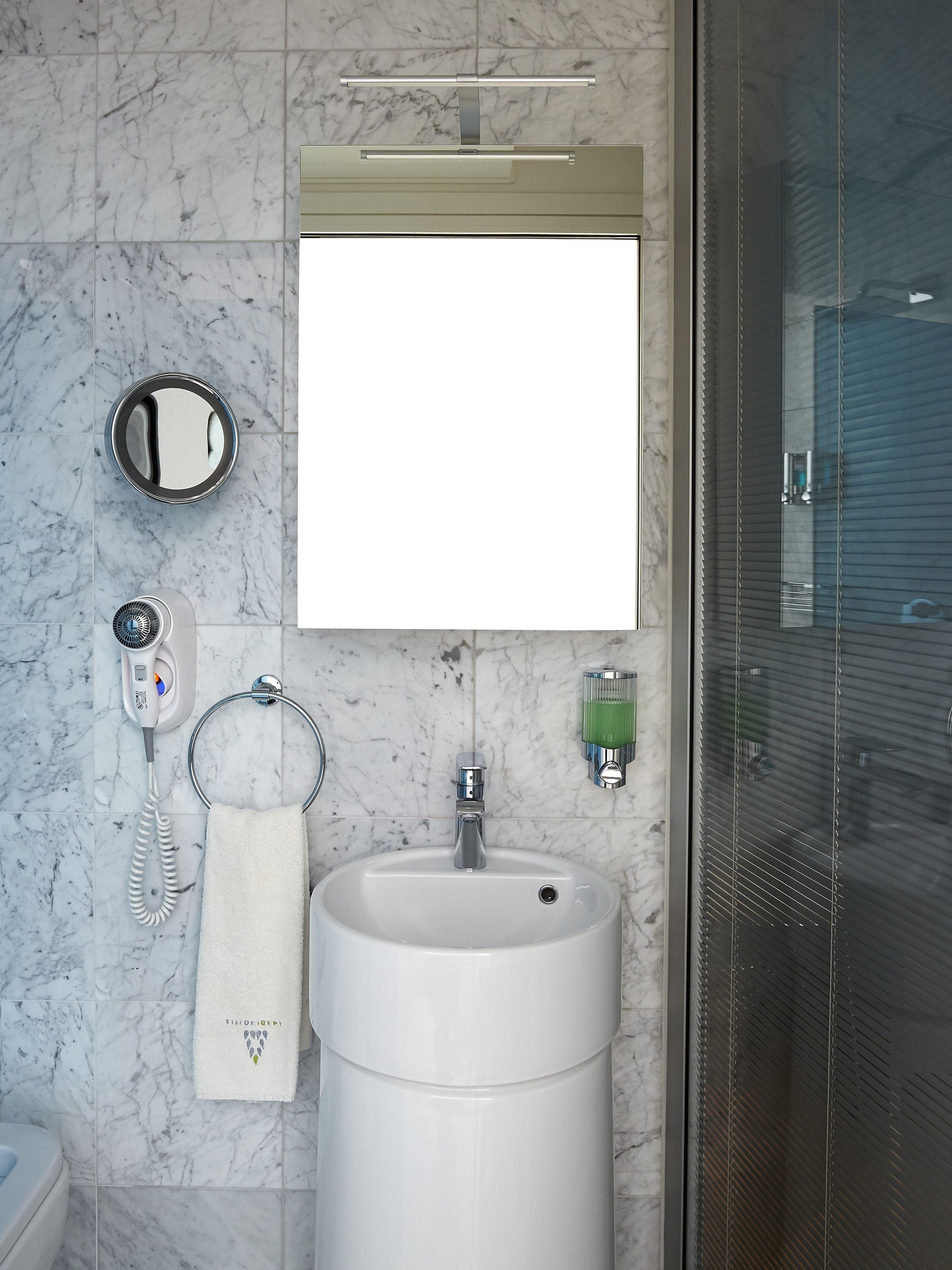 A bathroom with a marble floor, a white sink, a towel ring, a hairdryer, a mirror, and a glass shower enclosure.