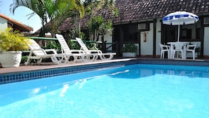 A swimming pool with white lounge chairs and a table with chairs, surrounded by potted plants and a building with a tiled roof. - Barla Inn Suites (Búzios)