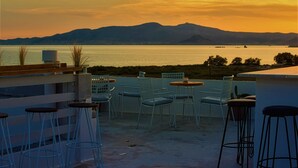 A terrace with outdoor seating and a view of a body of water and mountains during sunset. - Studios Vrettos Beachfront Hotel (Naxos)
