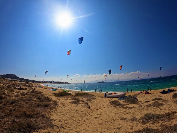 A beach with multiple kites flying in the sky, a clear blue ocean, and a sandy shore with some vegetation. - Studios Vrettos Beachfront Hotel (Naxos)