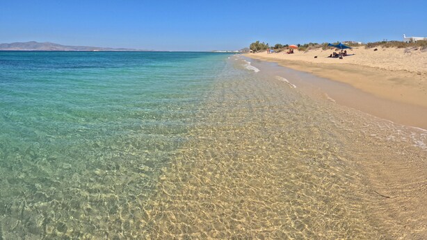 A beach with clear shallow waters, sandy shore, and a few people and umbrellas. - Studios Vrettos Beachfront Hotel (Naxos)