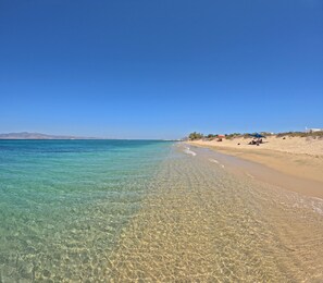 A beach with clear shallow waters, sandy shore, and a few people and umbrellas. - Studios Vrettos Beachfront Hotel (Naxos)