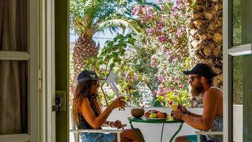 An open door leading to a patio with two people dining at a table, surrounded by greenery and a stone wall.