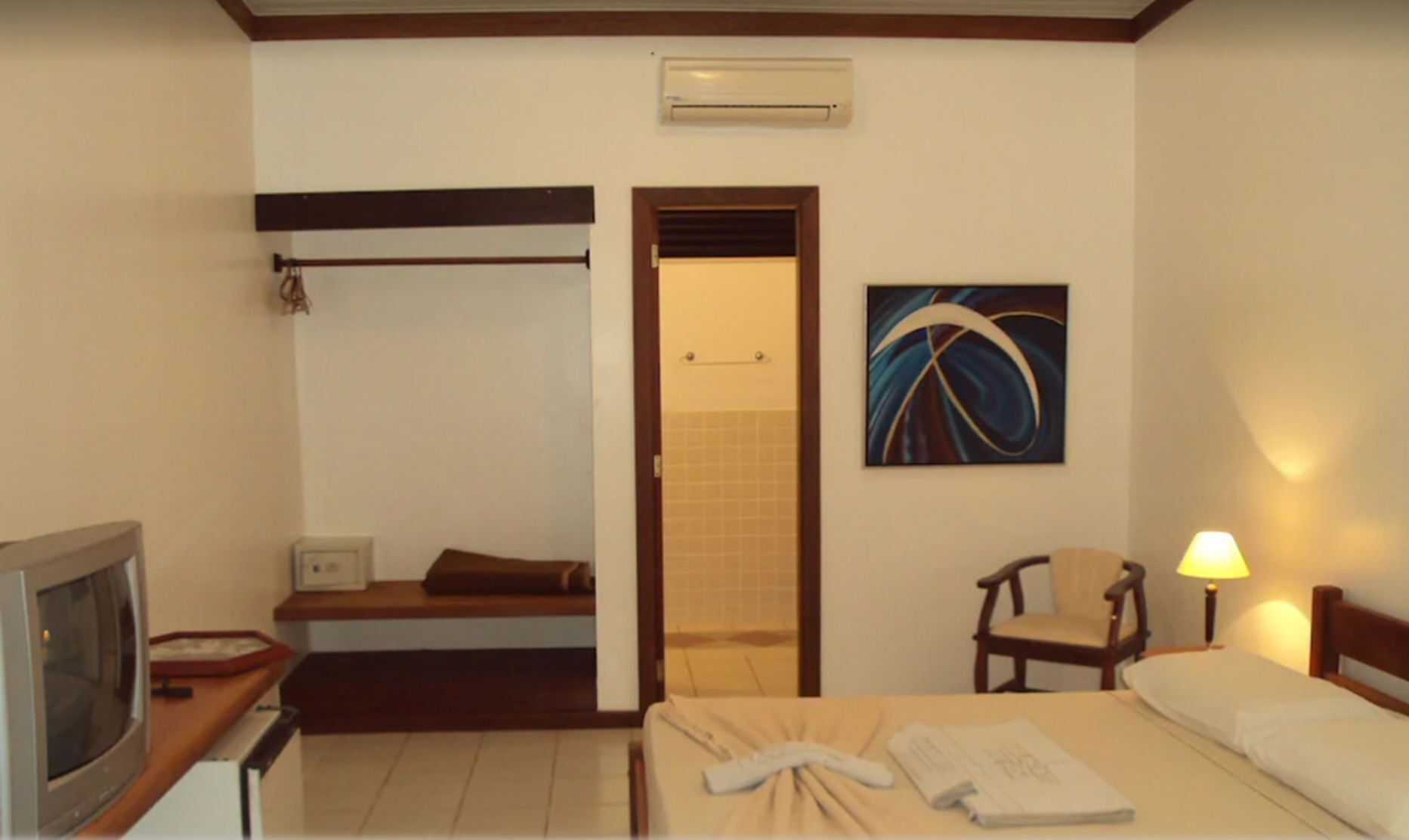 A hotel room with a bed, a chair, a desk with a television, a bathroom with a towel rack, and a painting on the wall.