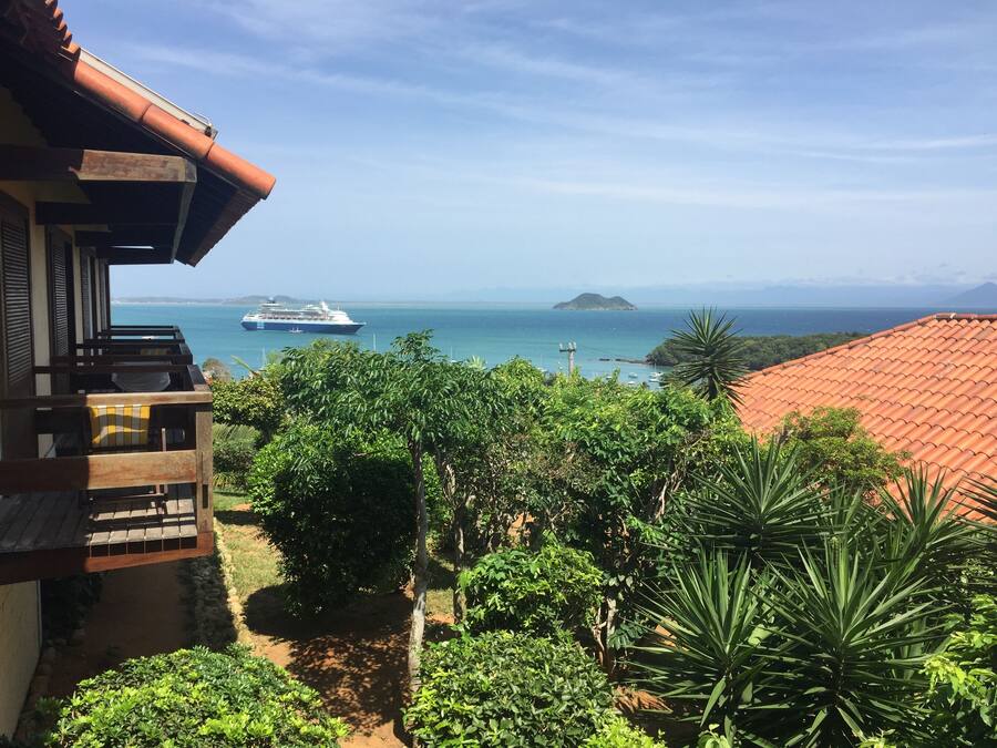 A coastal view with a cruise ship in the distance, lush greenery, and a building with a tiled roof.