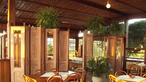 An indoor dining area with wooden tables and chairs, potted plants, and large windows allowing natural light.