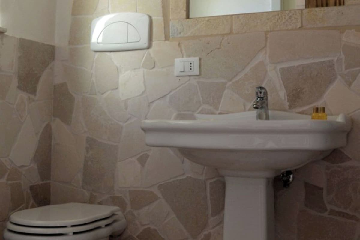 Une salle de bain avec un mur en pierre, un lavabo sur colonne blanc et des toilettes.