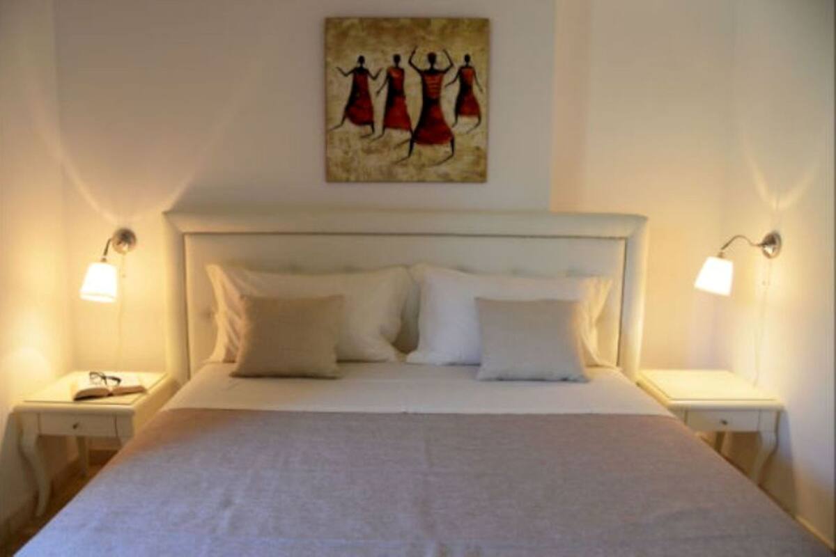 Ein ordentlich bezogenes Bett mit weißen Leinen und Kissen, zwei Nachttische mit Lampen und ein Wandbild mit drei Personen.