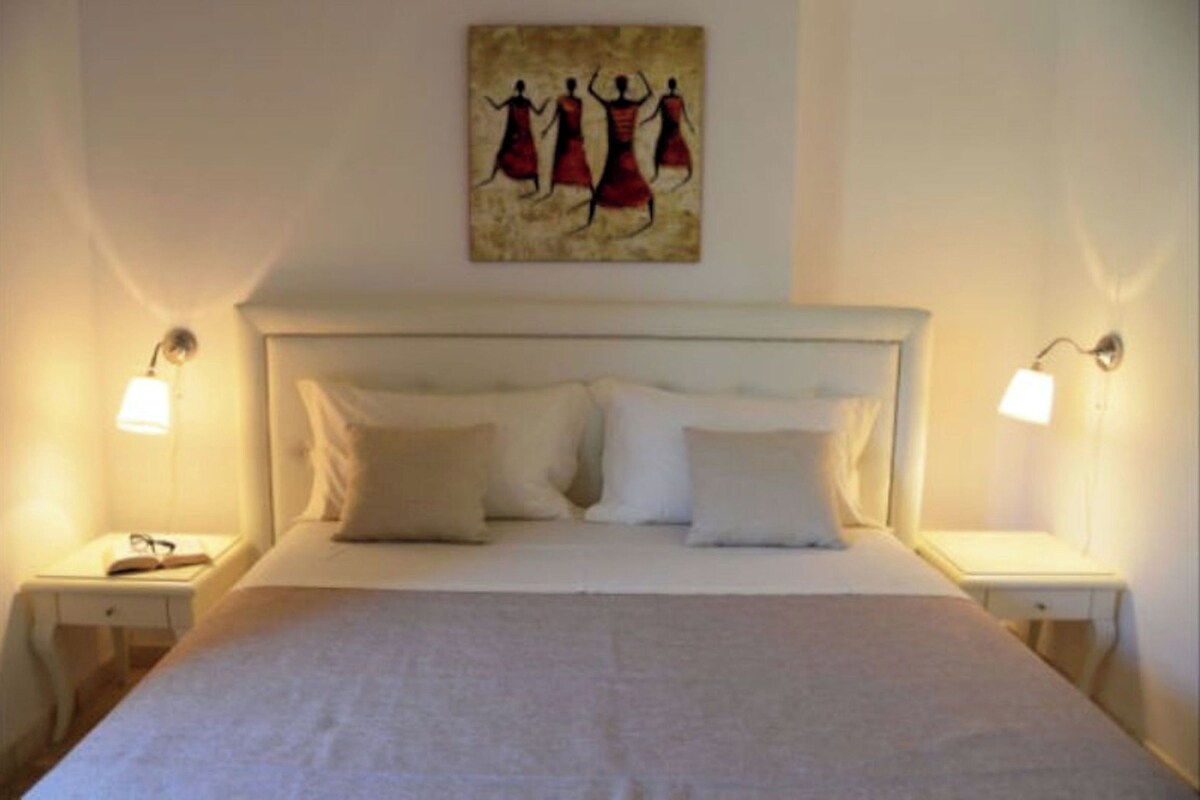 Ein ordentlich bezogenes Bett mit weißen Leinen und Kissen, zwei Nachttische mit Lampen und ein an der Wand hängendes Kunstwerk mit der Darstellung von drei Personen.
