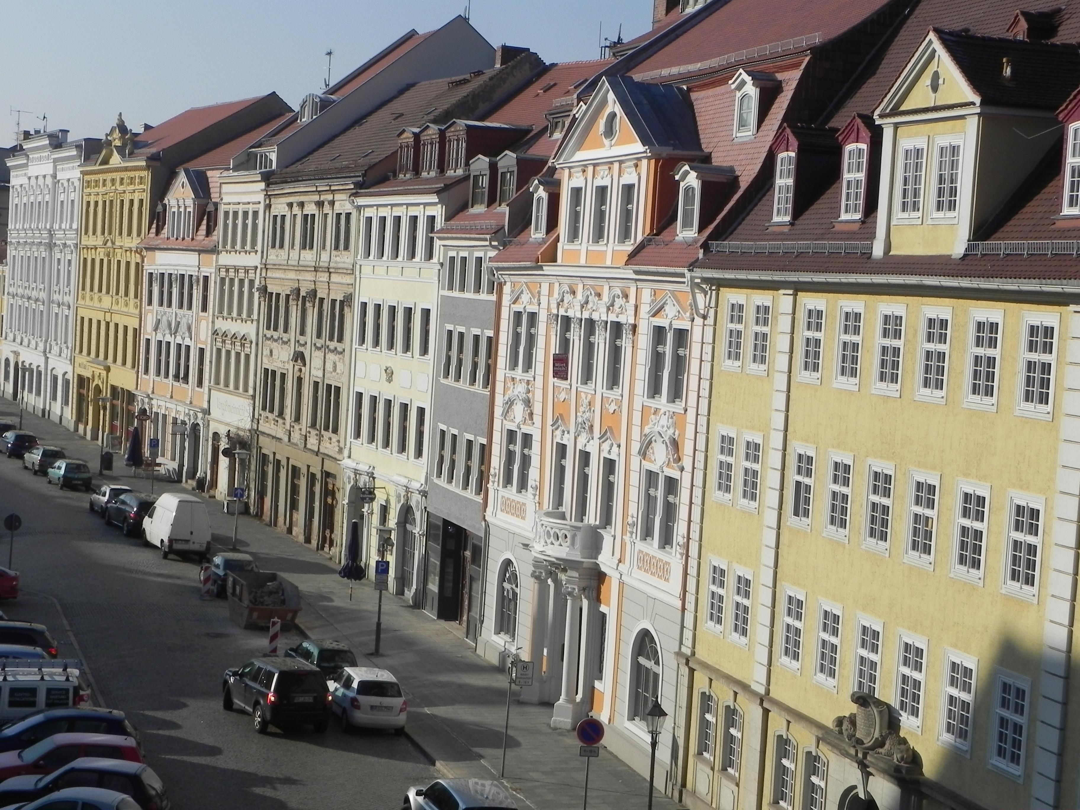 A row of historic buildings with uniform architectural styles, a street with parked cars, and a clear sky.