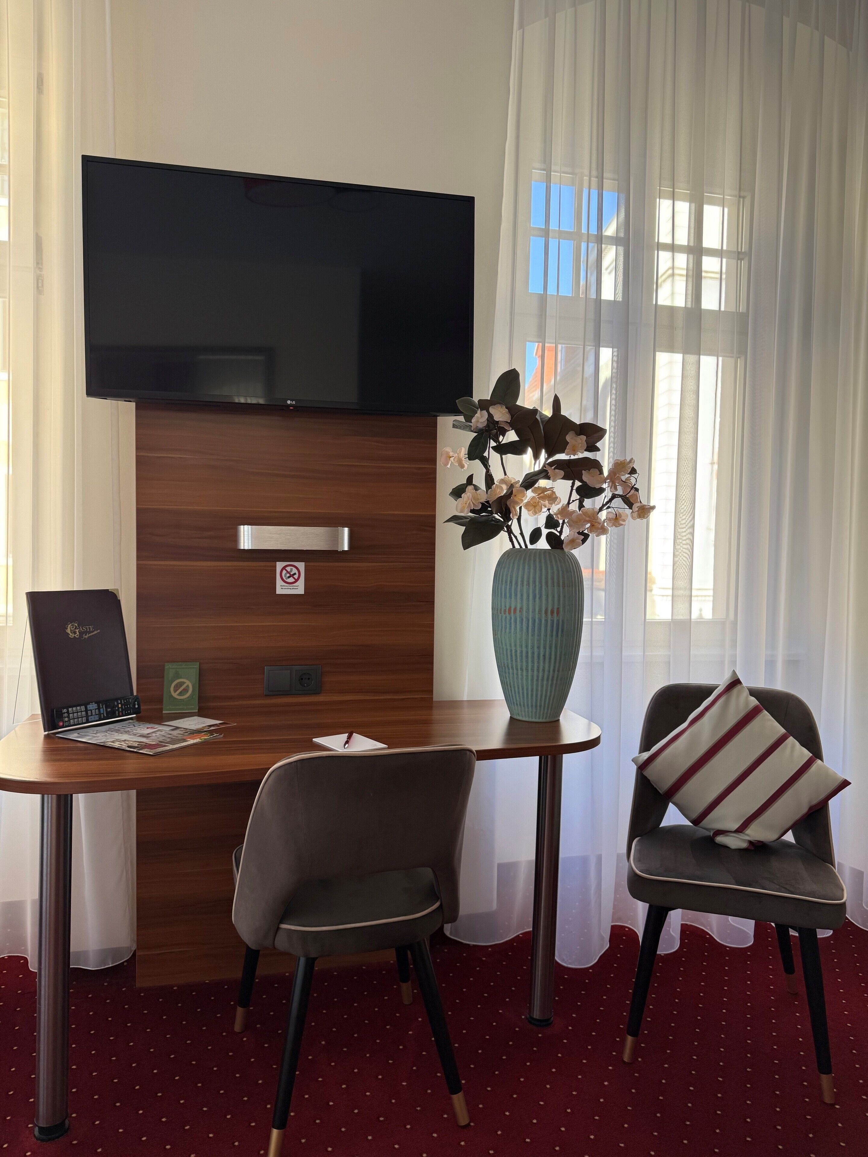 A hotel room with a flat-screen TV mounted on the wall, a wooden desk with a laptop, a vase with flowers, two chairs, and a red carpeted floor.