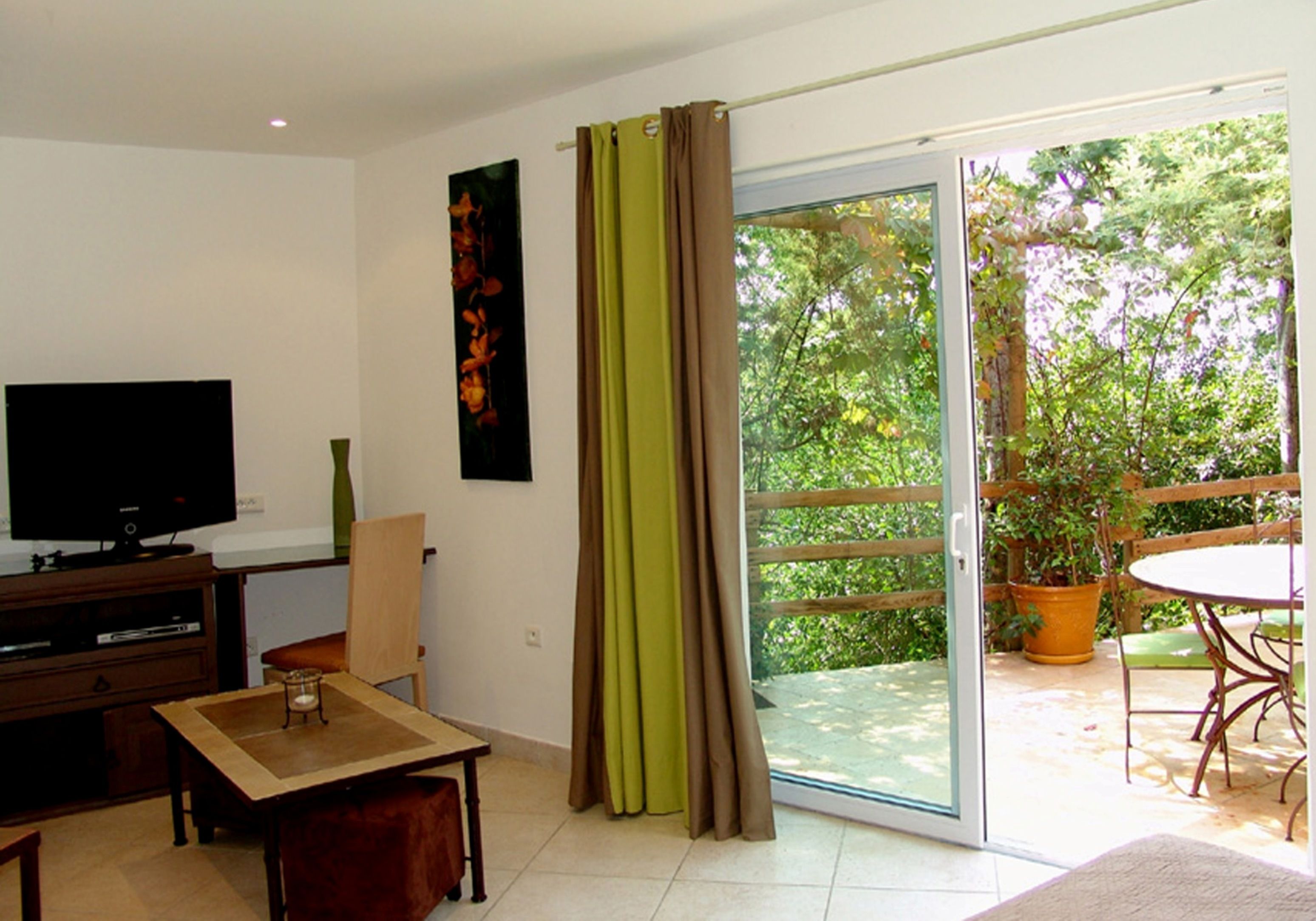 A living room with a television, a table, and a chair. There is a sliding glass door leading to an outdoor area with a table and chairs.