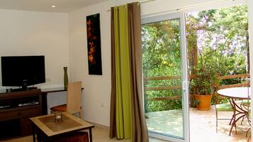 A living room with a television, a table, and a chair. There is a sliding glass door leading to an outdoor area with a table and chairs.