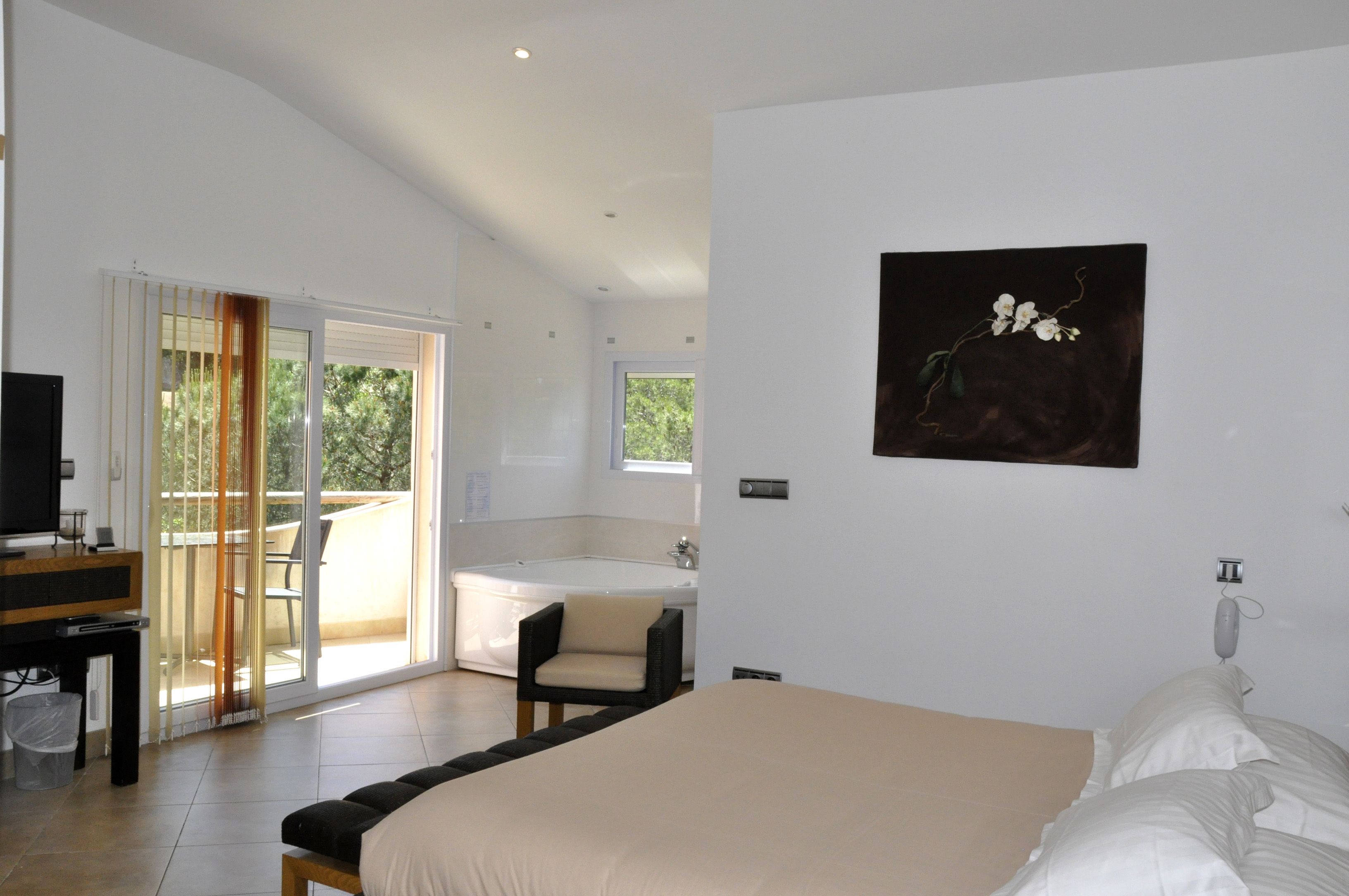 A modern bedroom with a large bed, a desk with a computer, a sliding glass door to a balcony, and a painting on the wall.