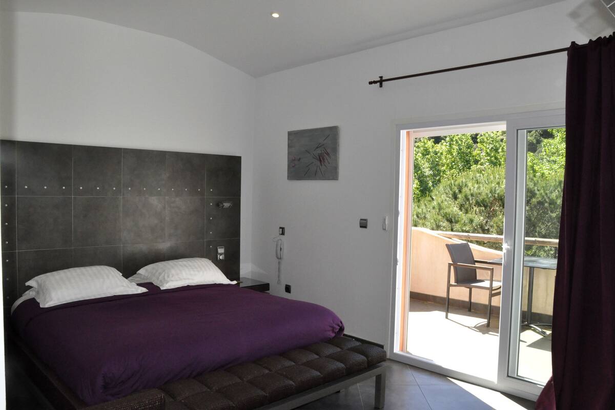 A bedroom with a large bed, a headboard, a painting on the wall, and a sliding glass door leading to an outdoor area with a chair.
