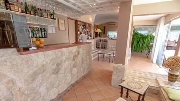 A bar area with a stone counter, various bottles, and glasses. There is a table with a vase and a cushion.