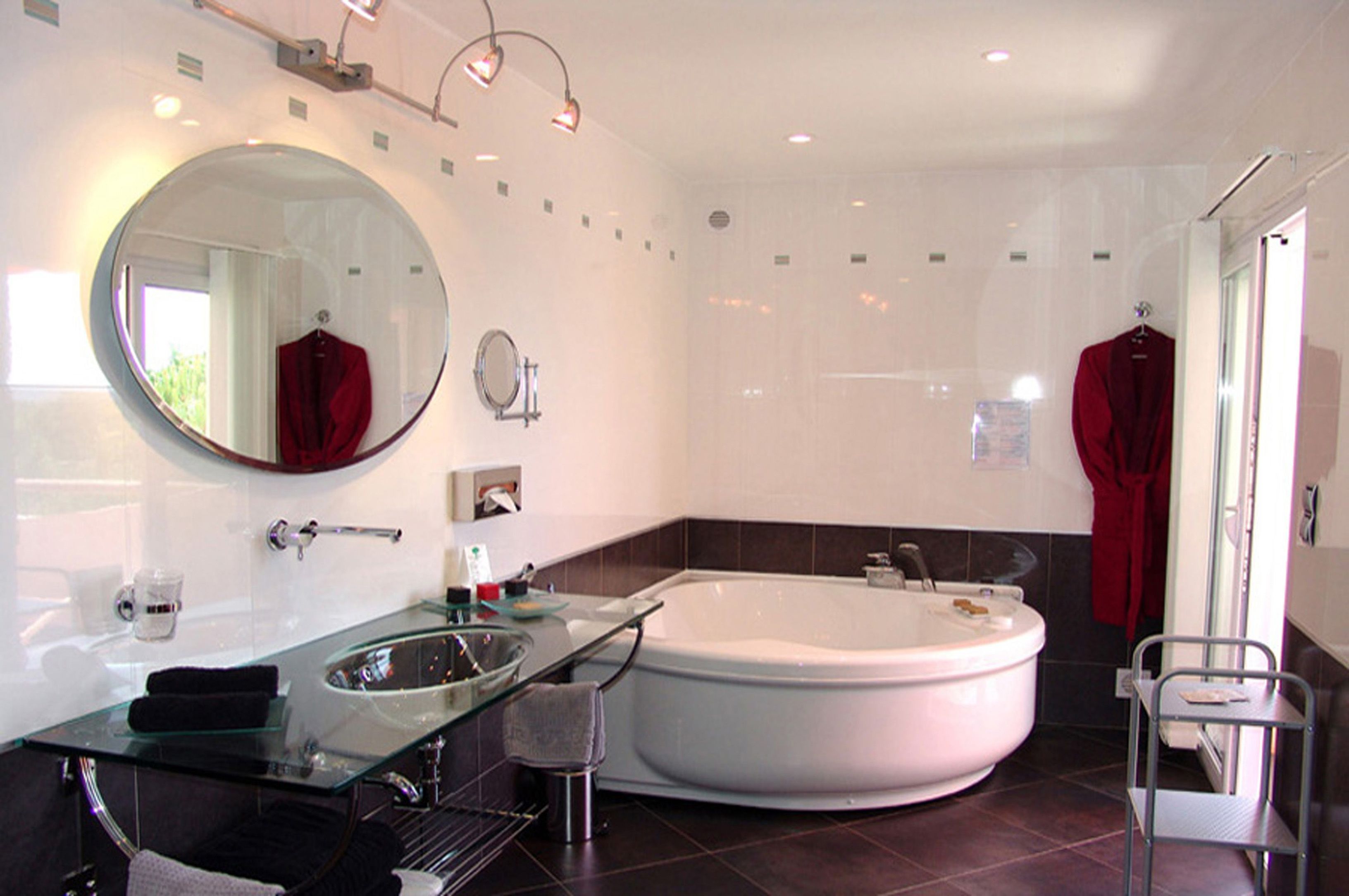 Een moderne badkamer met een glazen wastafel, een grote ronde spiegel, een vrijstaand bad en een rode handdoek aan een rek.