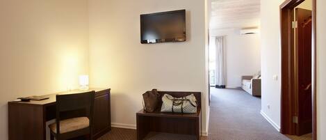 Habitación de hotel con escritorio, silla y un banco con maletas. Hay un televisor montado en la pared y una puerta que da a otra habitación.