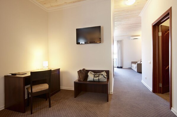 Habitación de hotel con escritorio, silla y un banco con maletas. Hay un televisor montado en la pared y una puerta que da a otra habitación.