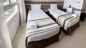 Een hotelkamer met twee bedden, een raam met gordijnen, een nachtkastje met een lamp en uitzicht op de stad.