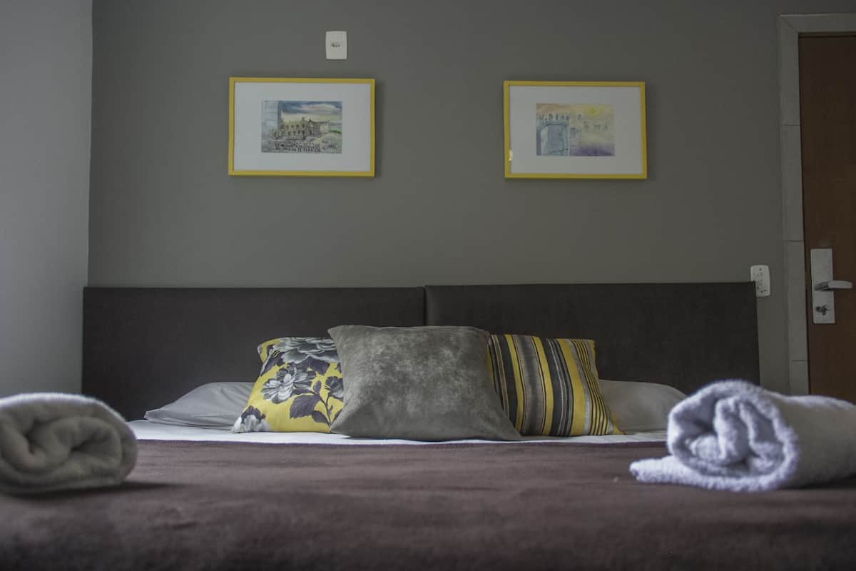 Ein ordentlich bezogenes Bett mit dunklem Kopfteil, zwei gerahmte Bilder an der Wand und eine Tür auf der rechten Seite.