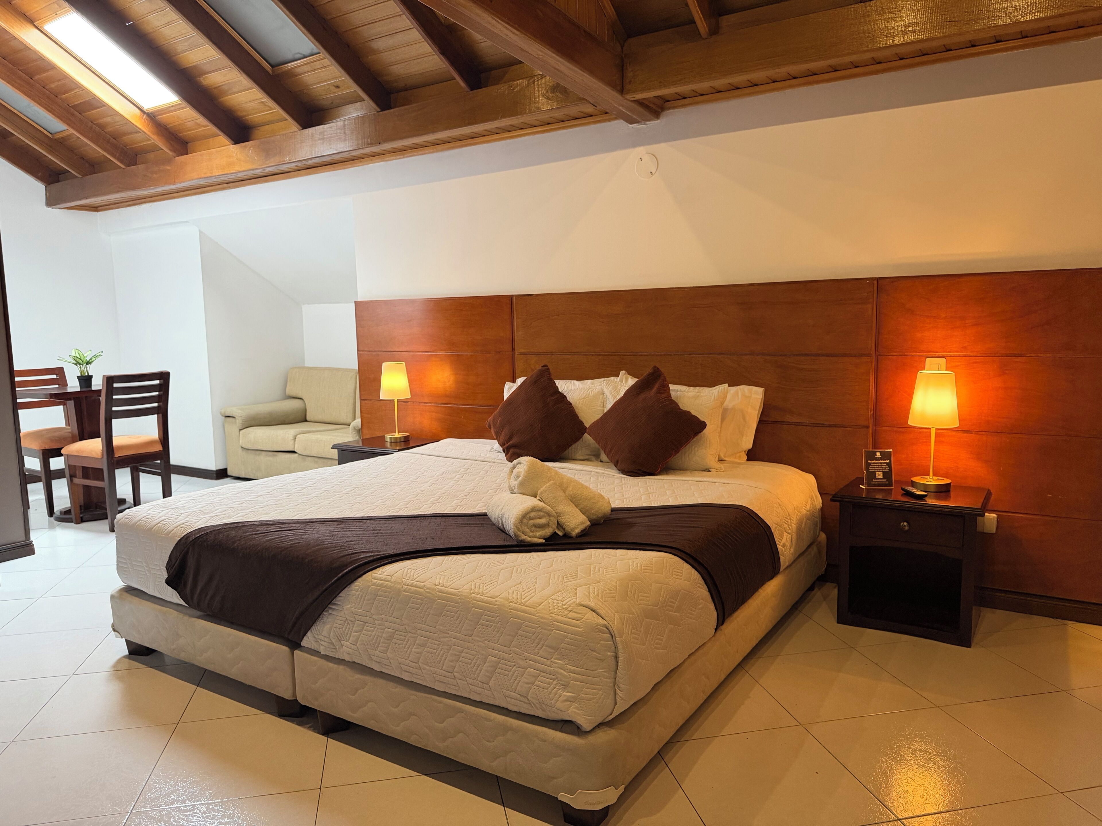 A hotel room with a large bed, wooden headboard, two bedside lamps, a small table, and a chair.