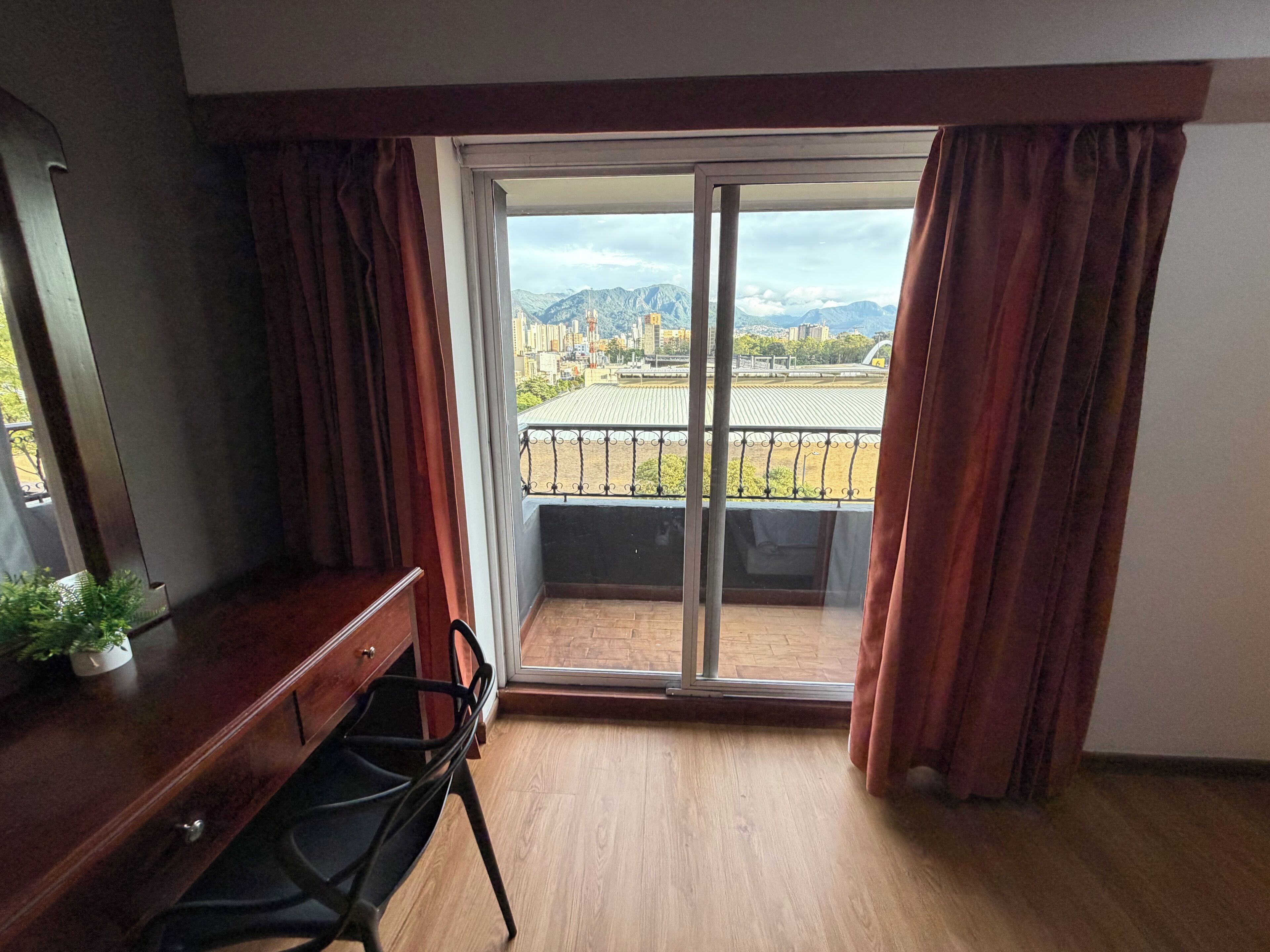 A room with a wooden dresser, a chair, a sliding glass door leading to a balcony with a view of mountains and buildings.