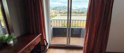 A room with a wooden dresser, a chair, a sliding glass door leading to a balcony with a view of mountains and buildings.