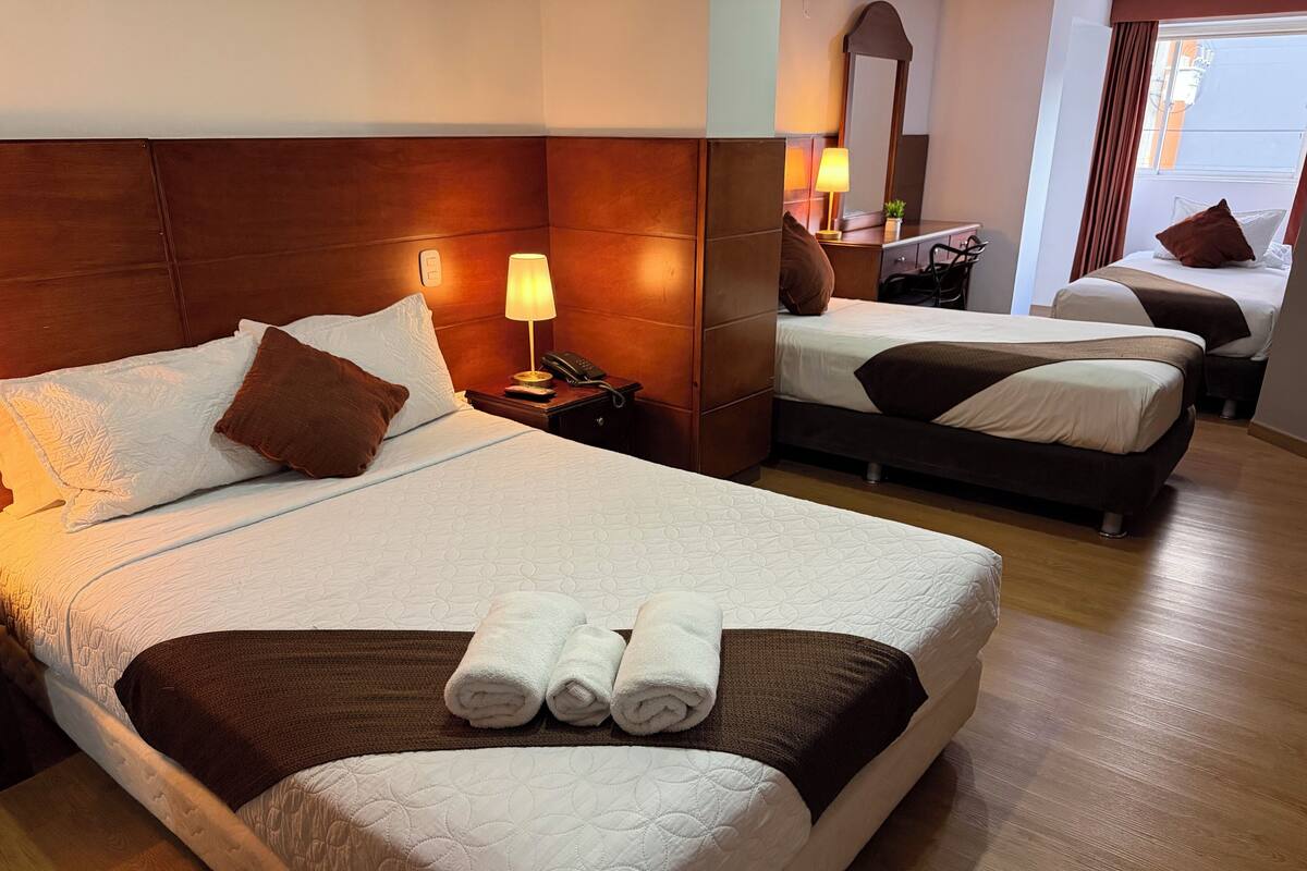 A hotel room with a large bed, a smaller bed, a nightstand with a lamp, a mirror, and a window with curtains.