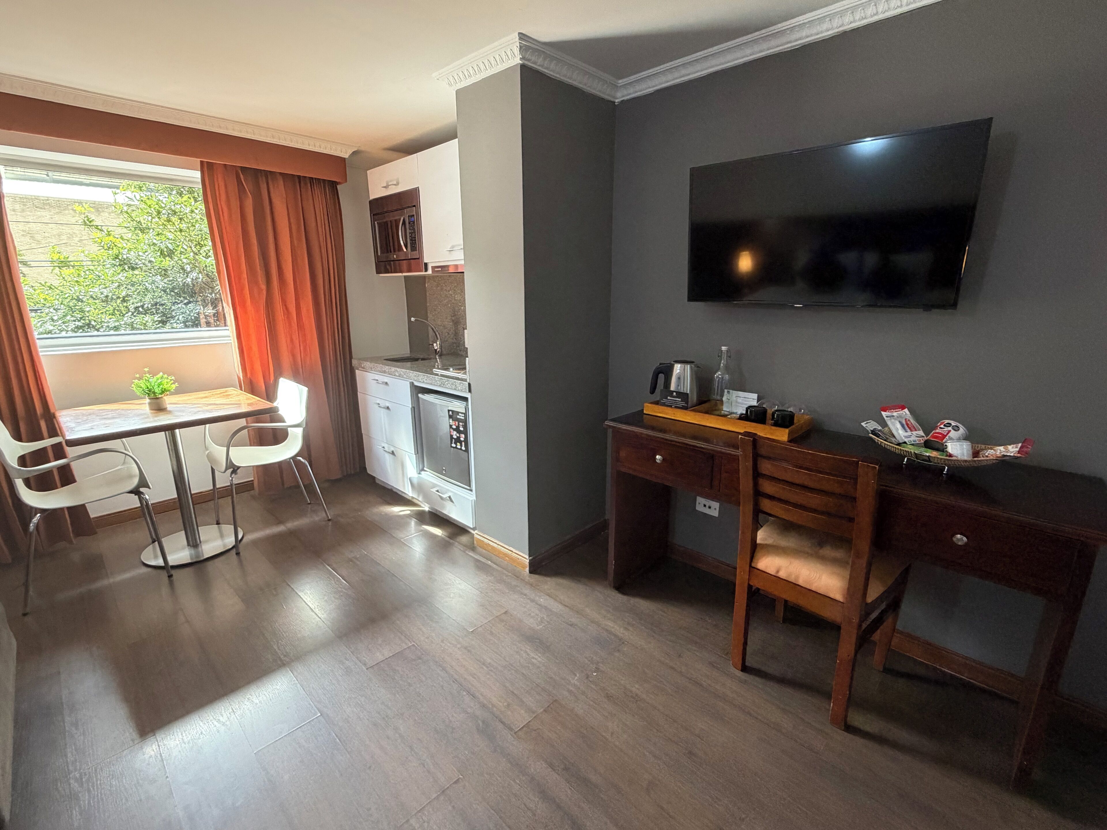 A compact hotel room with a kitchenette, a desk with a chair, a TV mounted on the wall, and a window with curtains.